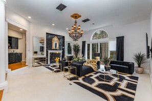 A spacious living room with a black and gold color scheme, a large chandelier, and a view of the outdoors through large windows.