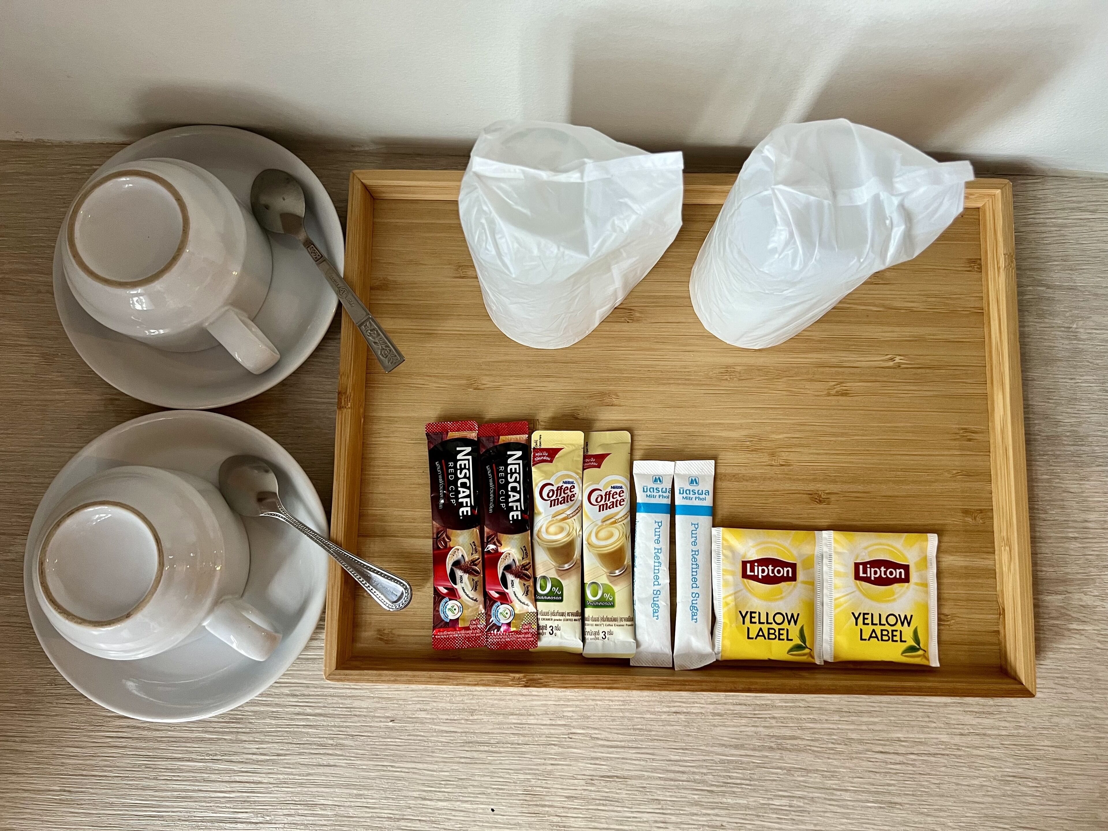Una bandeja con dos tazas de café, una cuchara y varios pods de café, incluyendo Nescafe Red Cup y Lipton Yellow Label.