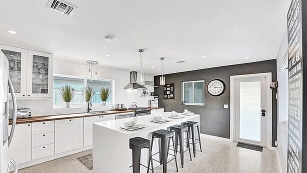 A modern kitchen with white cabinetry, a central island with bar stools, a large window, and a clock on the wall. - Beach Bliss: Heated Pool, BBQ & Tropical Vibes 🌊 (Oakland Park)