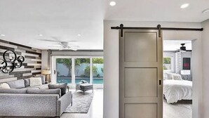 A modern living room with a grey sofa, a coffee table, and a decorative wall. There is a sliding door leading to a bedroom with a bed and a bathroom with a shower and a toilet. - Beach Bliss: Heated Pool, BBQ & Tropical Vibes 🌊 (Oakland Park)