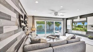 A modern living room with a gray sectional sofa, a ceiling fan, and a view of a swimming pool through large sliding glass doors. - Beach Bliss: Heated Pool, BBQ & Tropical Vibes 🌊 (Oakland Park)
