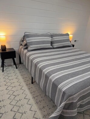 A bed with a striped bedspread and pillows, a nightstand with a lamp, and a patterned rug.