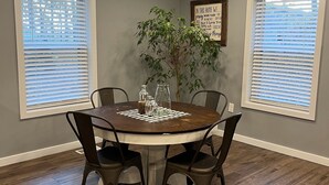 A dining room with a round table, four chairs, a potted plant, and two windows with blinds.