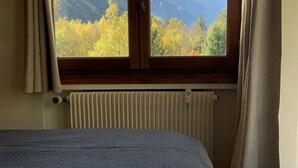 Ein Schlafzimmer mit Bett, einem Fenster mit Vorhängen und Blick auf Berge und Herbstbäume.