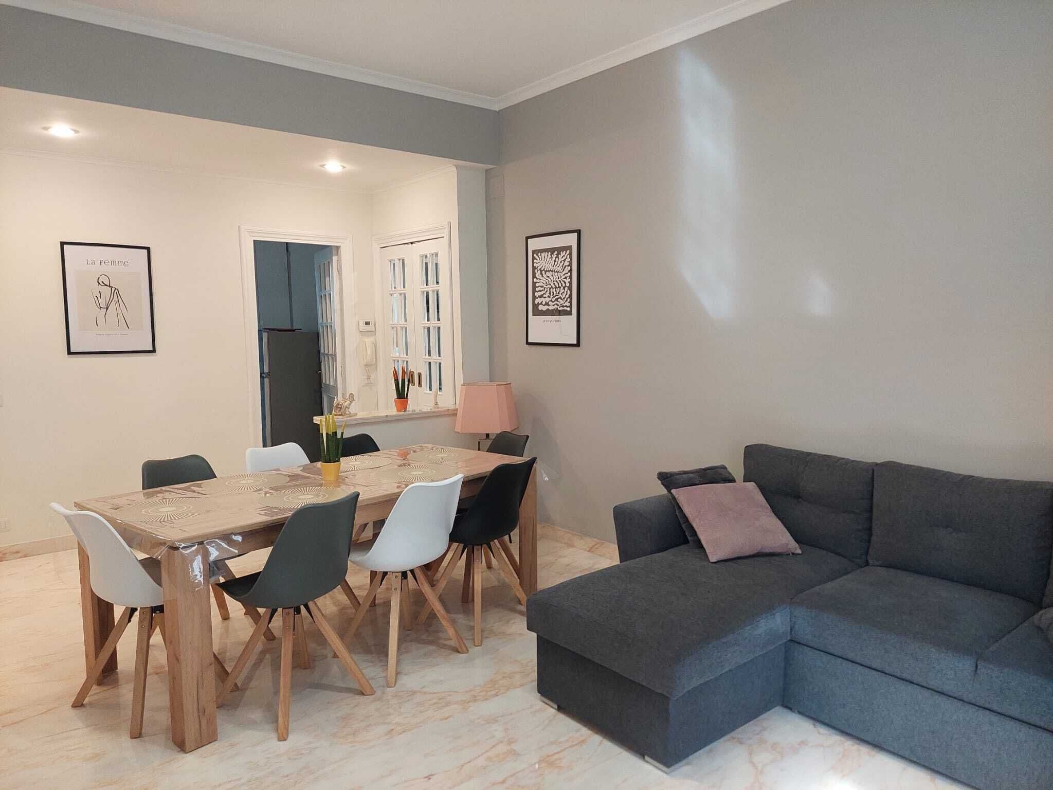 A modern living room with a grey sofa, a dining table with chairs, and framed artwork on the walls.