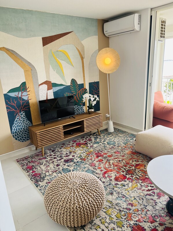 A living room with a flat-screen TV, a colorful rug, a round ottoman, and a wall mural. - CHIC SUITE WITH PANORAMIC OCEAN VIEWS. BEACH & SWIMMING POOL IN MONT VERNON, EASTERN BAY (Collectivité de Saint-Martin)