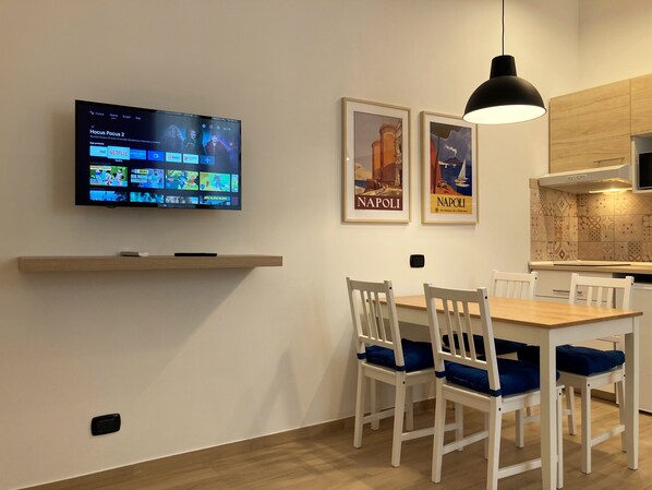 A modern dining area with a TV displaying a streaming service, two framed posters, a wooden shelf, and a kitchen area with a hanging light. - The Blue House 1 in the historic heart of Naples (Napoli)
