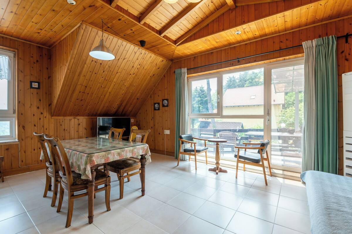 Een eetruimte met een glazen tafel, houten stoelen, een hanglamp en uitzicht door grote ramen.