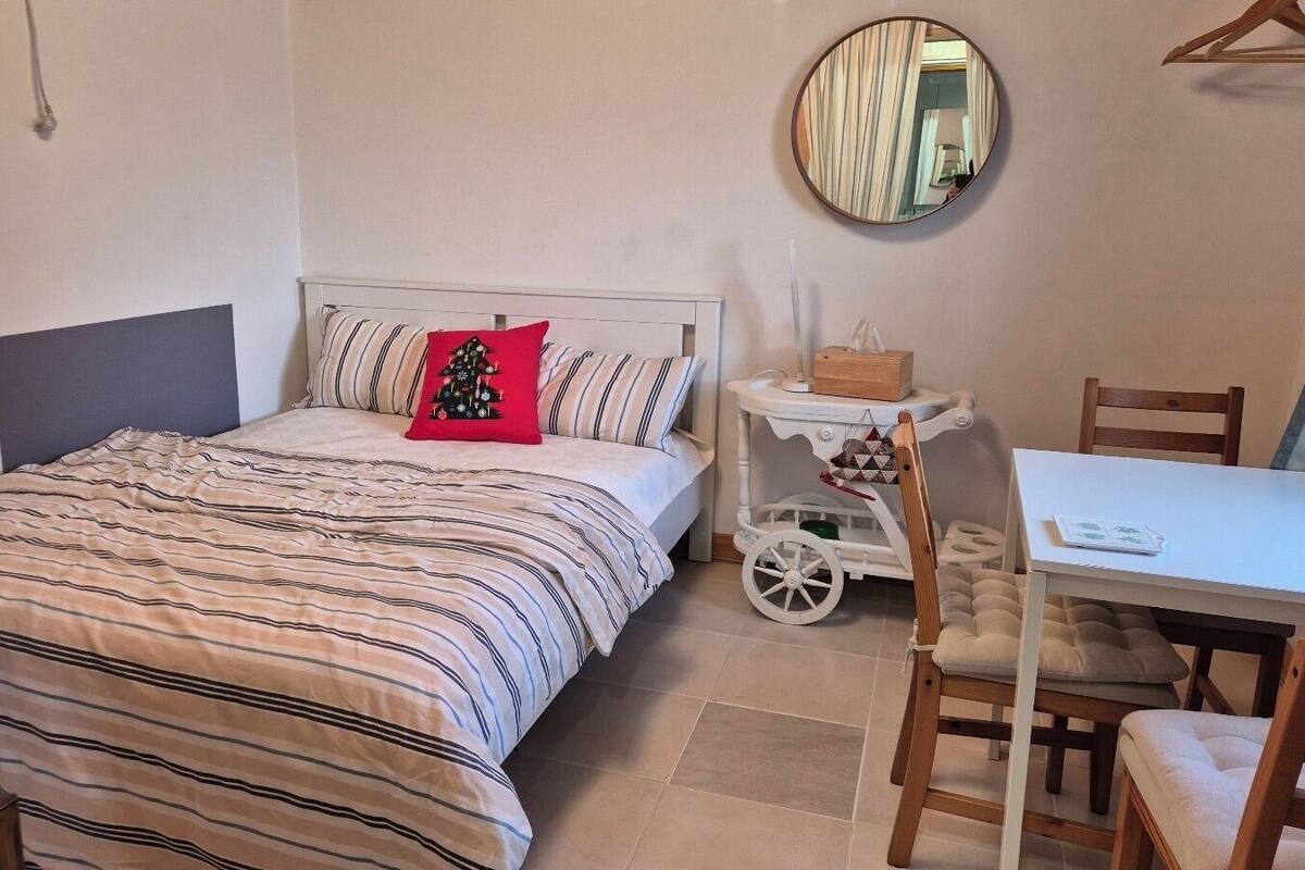 Een slaapkamer met een bed, een nachtkastje met een lamp, een kleine tafel met stoelen en een ronde spiegel aan de muur.