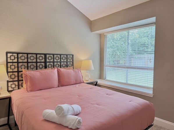 Habitación de hotel con cama, dos almohadas, mesita de noche con lámpara y ventana con persianas.