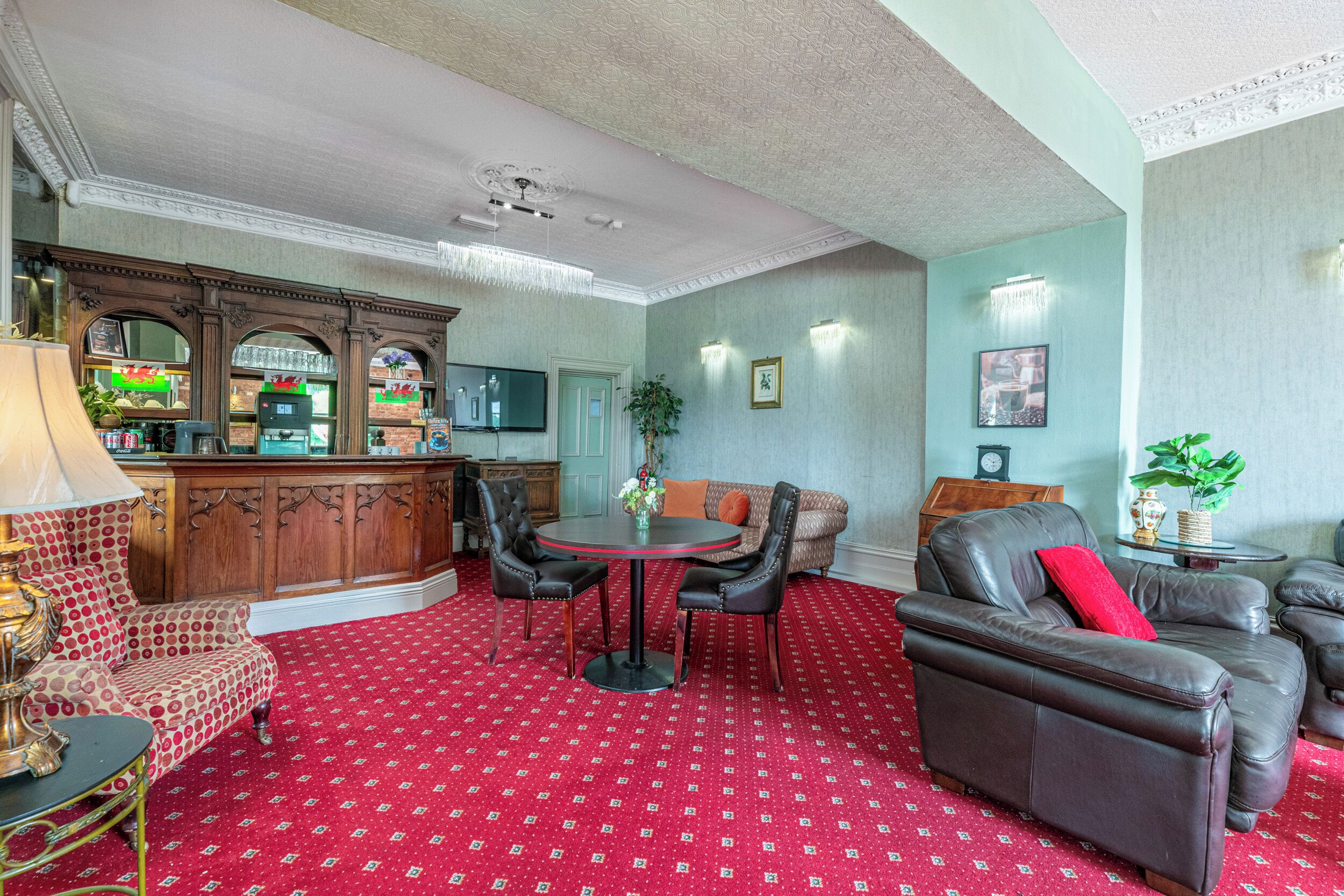 A room with a red patterned carpet, a round table with chairs, a sofa, a bar area with a television, and a potted plant.
