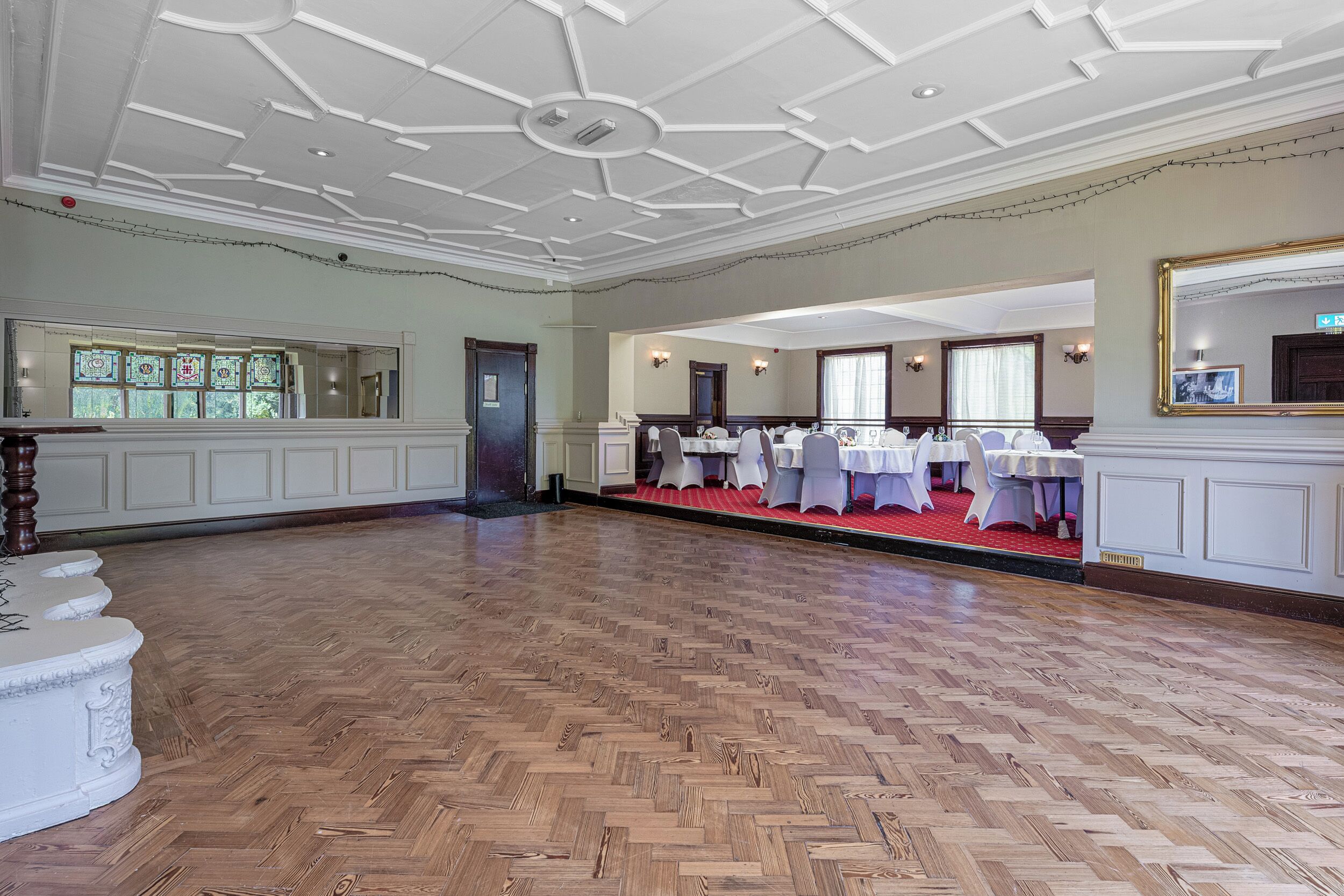 Ein geräumiger Saal mit Holzdielenboden, weißen Wänden und einer Decke mit geometrischem Muster.