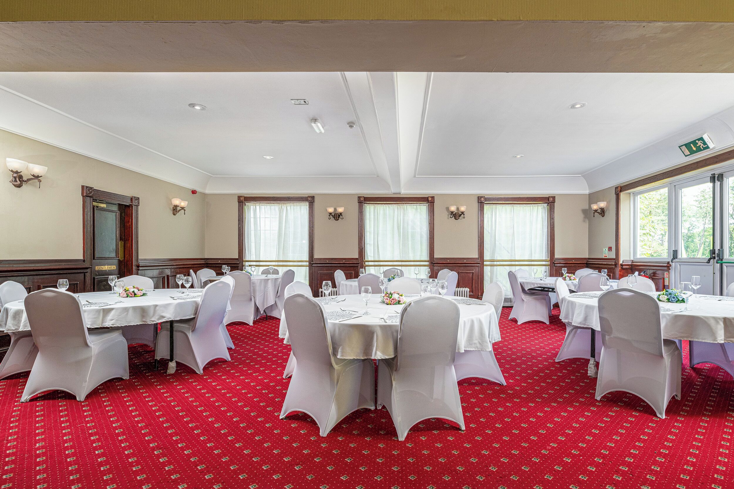 Un salón de banquetes con mesas redondas, sillas blancas y suelo cubierto de alfombra roja.