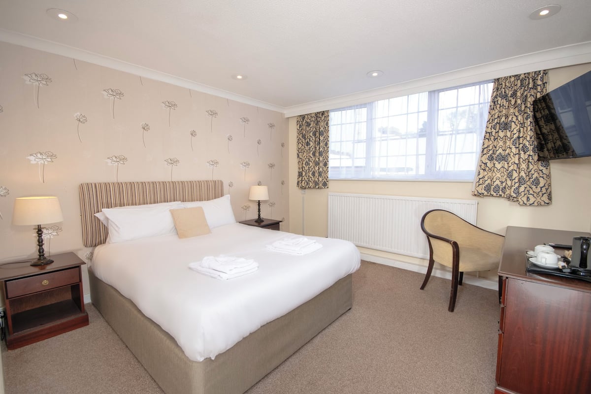 A hotel room with a large bed, two bedside tables with lamps, a wooden chair, a desk with a television, and a window with patterned curtains.