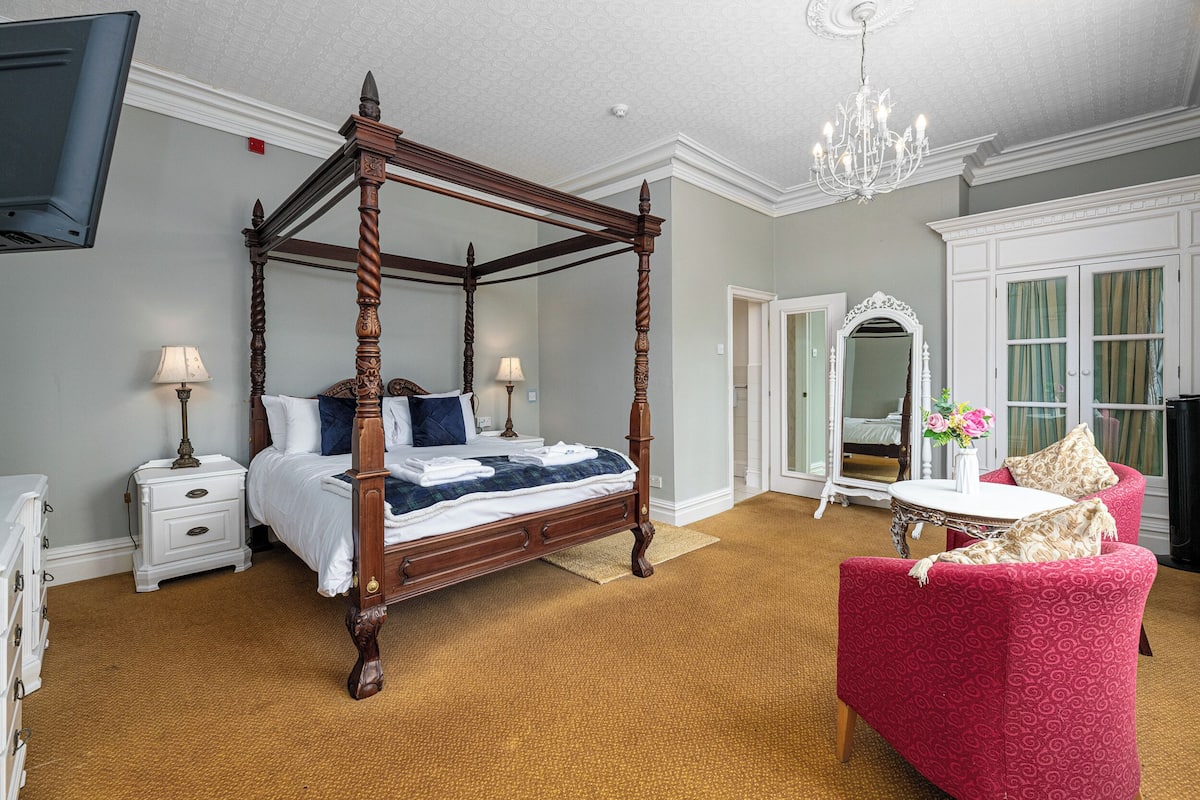 A four-poster bed with a wooden frame, a red upholstered chair, a white bedside table, and a large mirror.