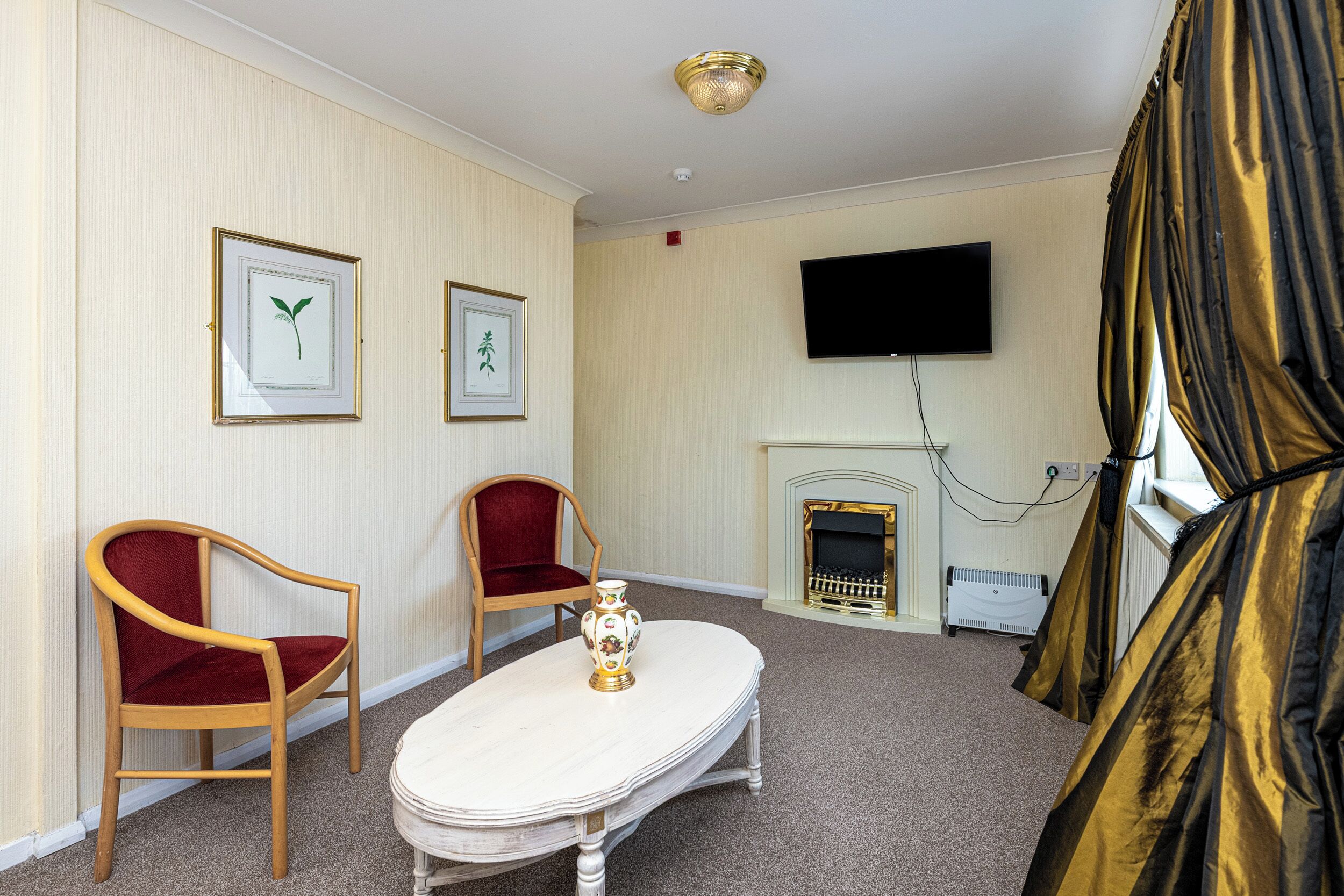A room with two wooden chairs, a cream-colored table, a flat-screen TV, a fireplace, and a window with curtains.
