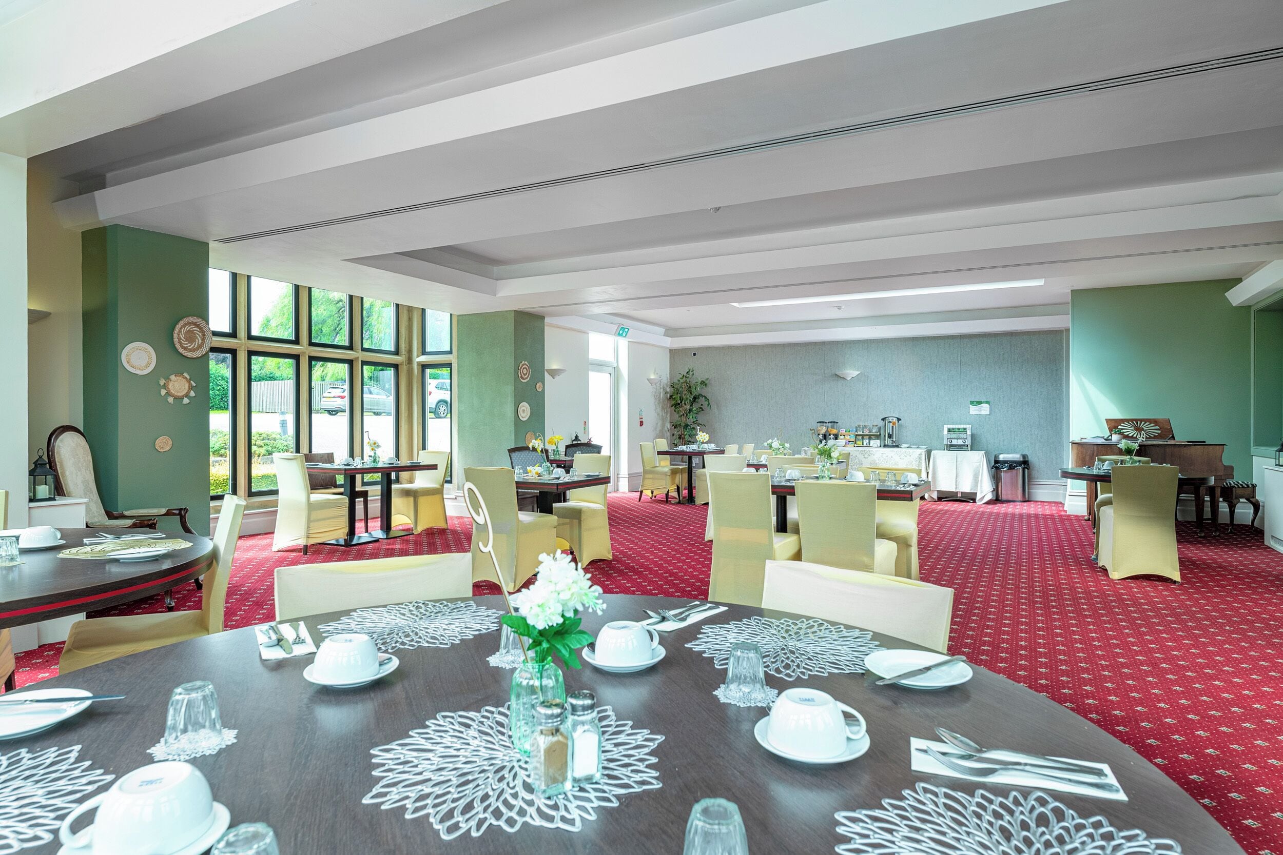 A dining area with round tables set for four, each with white tableware and a centerpiece. The room has green walls, large windows, and a red carpet.