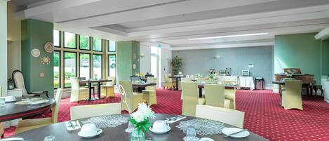 A dining area with round tables set for four, each with white tableware and a centerpiece. The room has green walls, large windows, and a red carpet.
