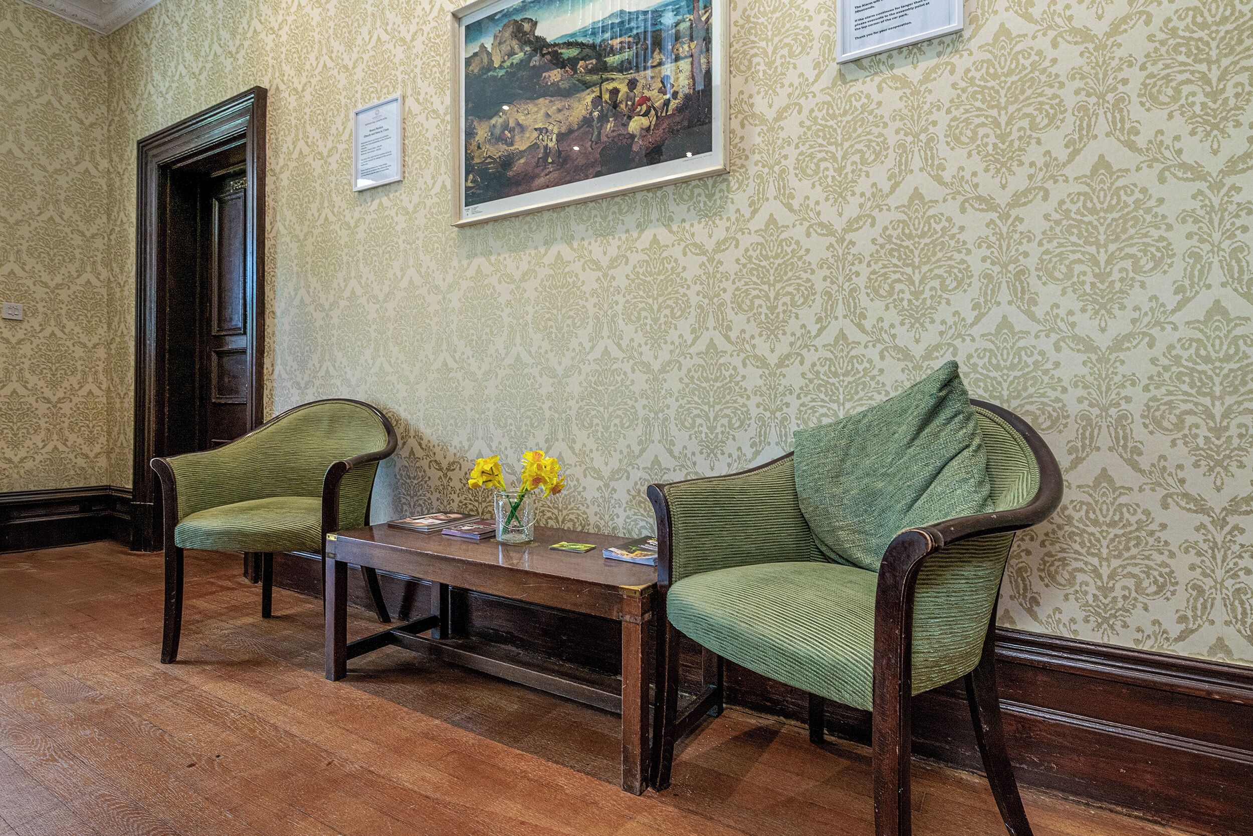 A room with two green upholstered chairs, a wooden table, and a framed painting on the wall.