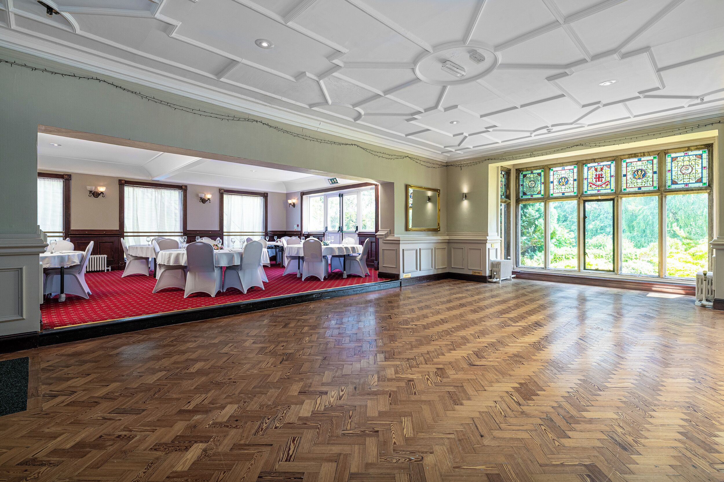 A spacious hall with a wooden parquet floor, white tables with chairs, and large windows with stained glass.