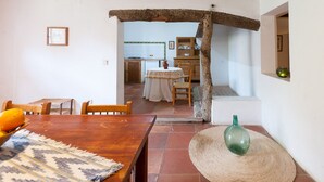 A rustic dining area with a wooden table, chairs, and a woven mat. A green bottle and a woven basket are on the floor. A doorway leads to a kitchen with a tiled backsplash and a table with a white tablecloth. - Family apartment Ca l'Amo (Sineu)