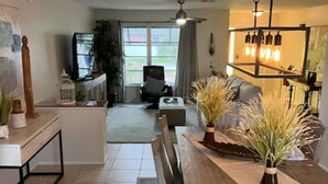 A modern living room with a wooden dining table, white chairs, and a sofa. There is a television and a plant in the background. - New on Market ! Beautiful family home close to beach, golf, tennis and shopping. (Venice)