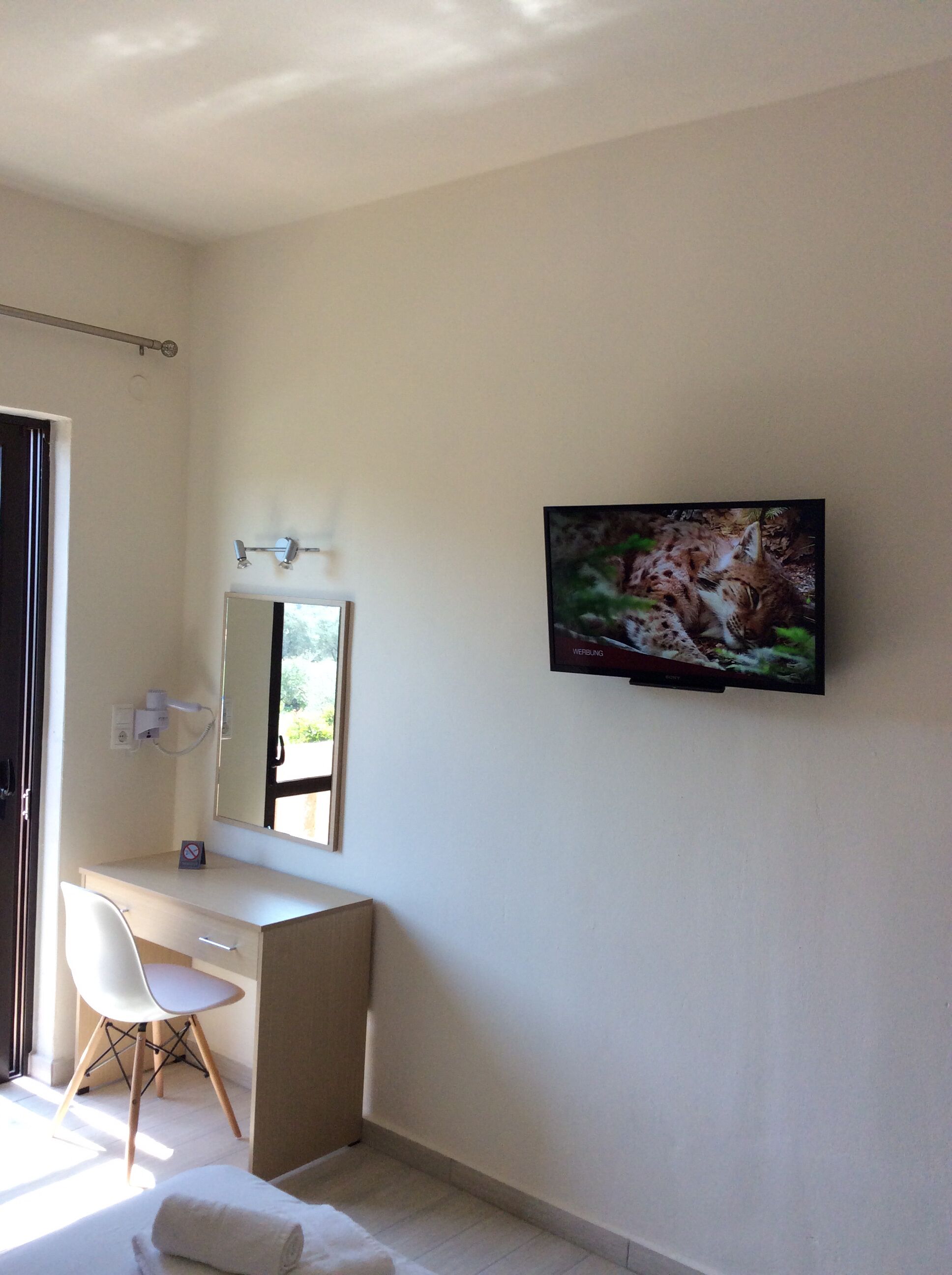 A modern bathroom with a white vanity, a mirror, a television mounted on the wall, and a window providing natural light.