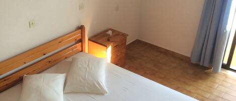 Una cama bien hecha con almohadas y toallas enrolladas, una mesita de noche de madera con lámpara, y una ventana con cortinas.
