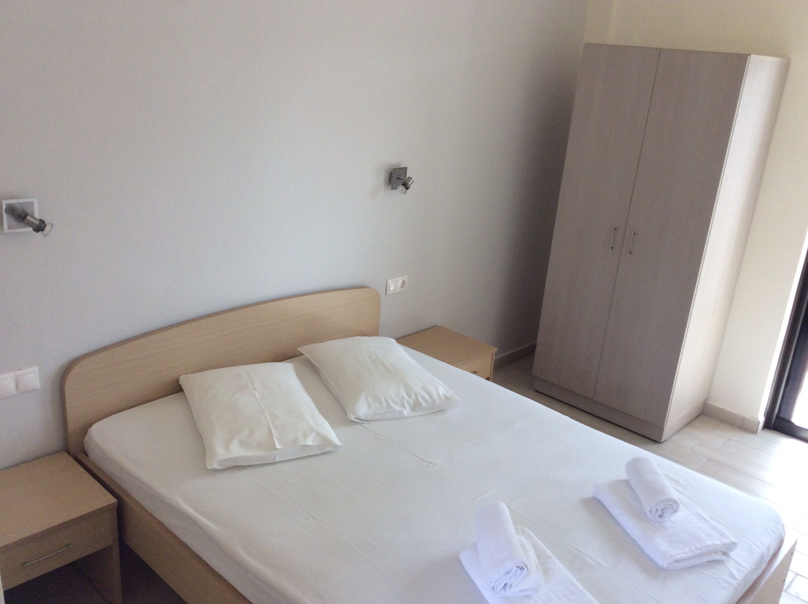 A neatly made bed with white linens and pillows, a wooden nightstand, a wardrobe, and a window providing natural light.