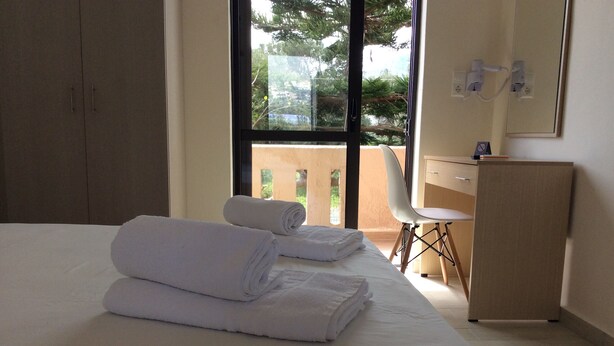 A hotel room with a bed, two rolled towels, a desk, a chair, and a view of greenery outside. - Elena Rooms (Agios Vasileios)