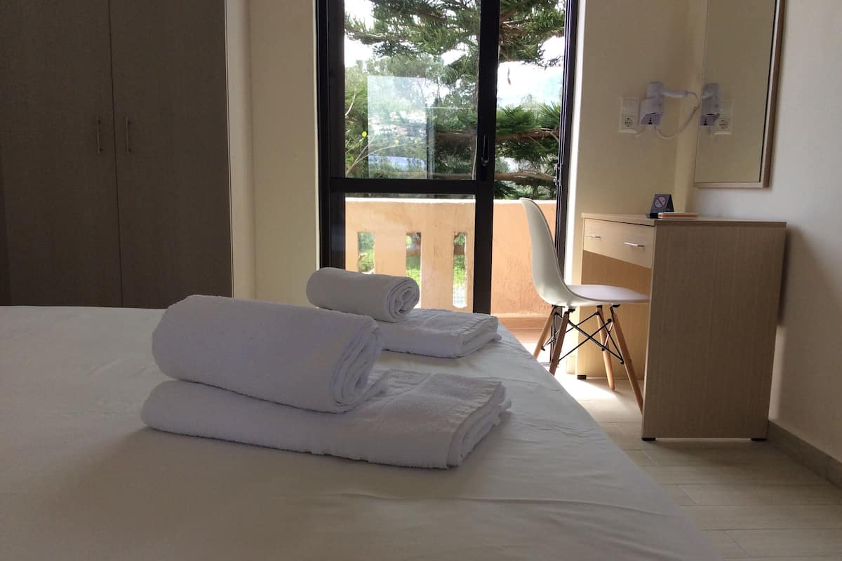 A hotel room with a bed, two rolled towels, a desk, a chair, and a view of greenery outside.