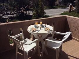 Une table ronde entourée de chaises blanches, sur un balcon, avec vue sur une route et la verdure.