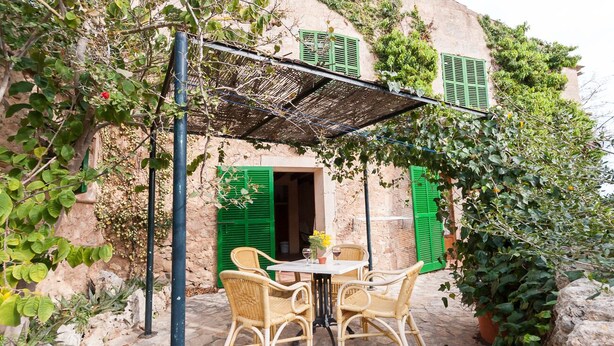 Un espacio de descanso rústico al aire libre con sillas de mimbre y una mesa, una pérgola con techo tejido, y un edificio con contraventanas verdes y plantas trepadoras.