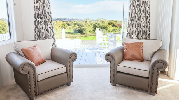 Two leather armchairs with cushions, a wooden coffee table, and a view of a garden and white chairs through a glass door. - Cherry Lodge (Newton Abbot)