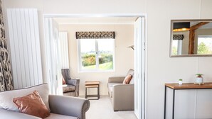 A modern living room with a sofa, armchair, and coffee table. There is a window with a view of greenery outside. - Cherry Lodge (Newton Abbot)