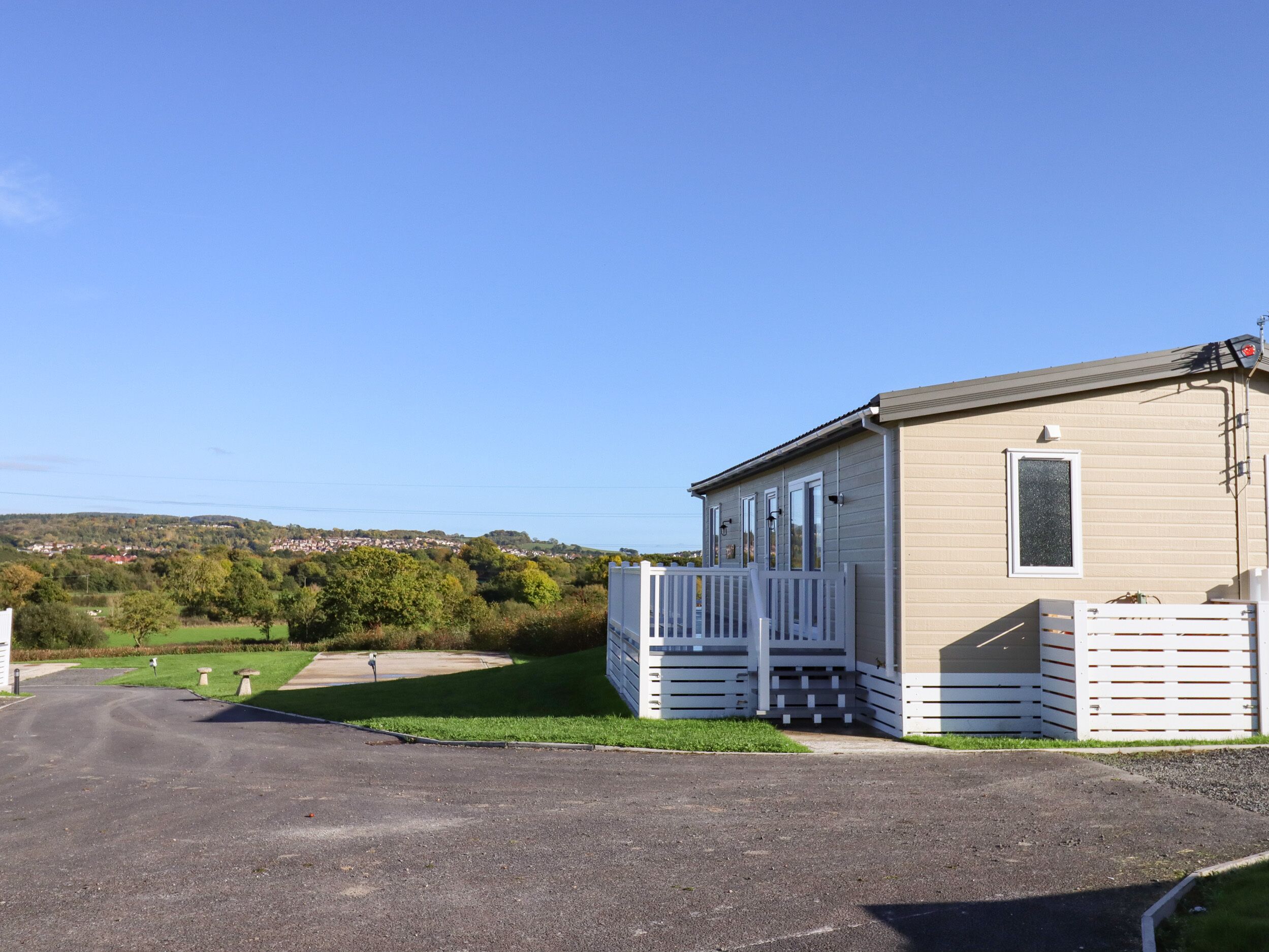 A single-story mobile home with a fenced porch and a small deck, set in a rural area with a clear sky.