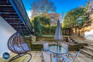 Relax in a serene outdoor space with a round table, hanging chair, and umbrella, surrounded by lush gardens and a multi-story building in the background.