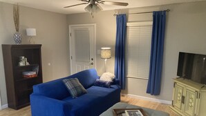 A living room with a blue sofa, a grey ottoman, a bookshelf, a floor lamp, and a television. - Fern’s Cottage New Construction! A cozy place to relax. (Atmore)