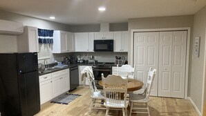 A compact kitchen with white cabinets, a black refrigerator, a microwave, and a dining area with two white chairs and a wooden table. - Fern’s Cottage New Construction! A cozy place to relax. (Atmore)