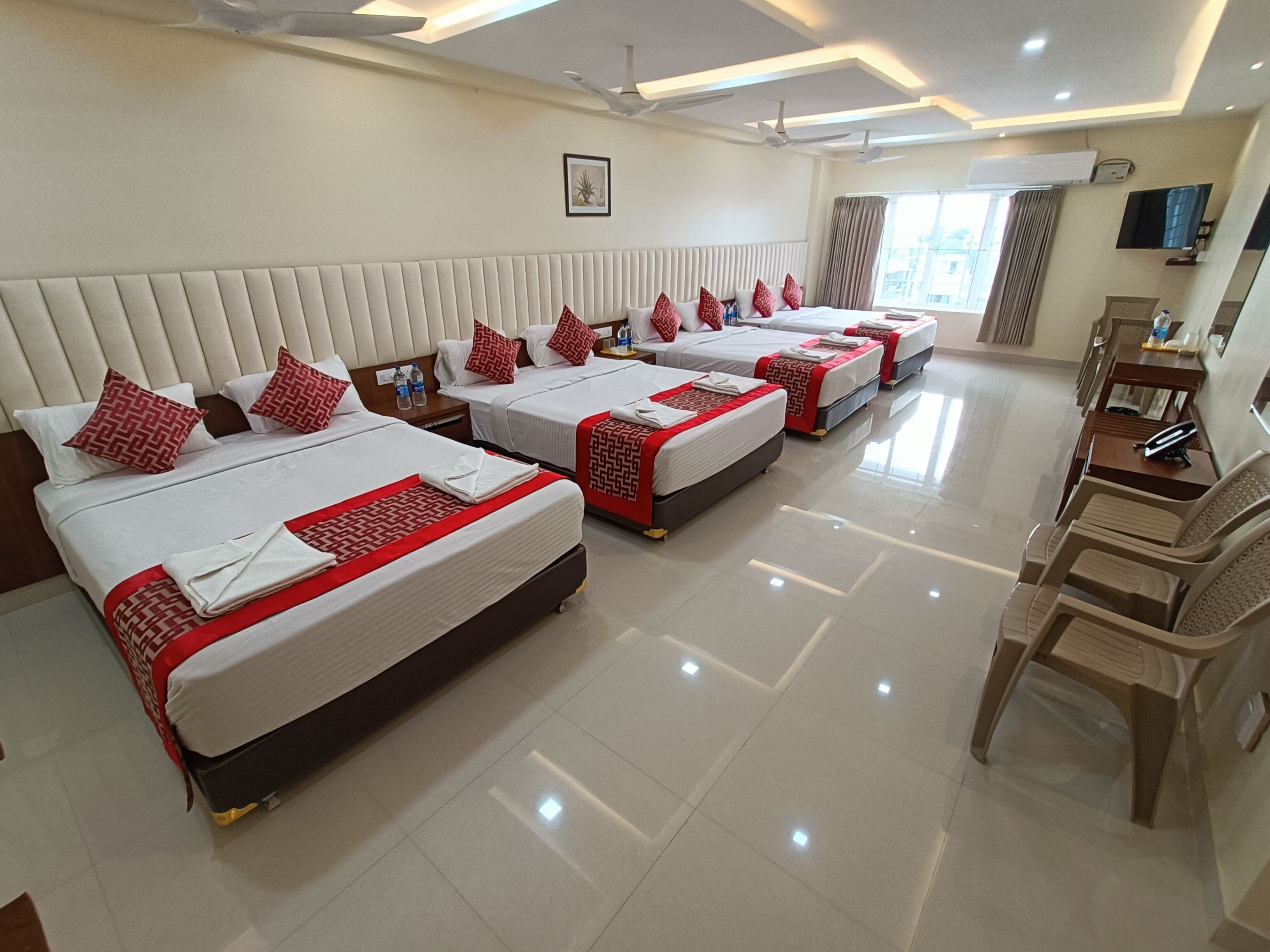 A hotel room with four beds, each with red and white bedding, a flat-screen TV, and a desk with a chair.