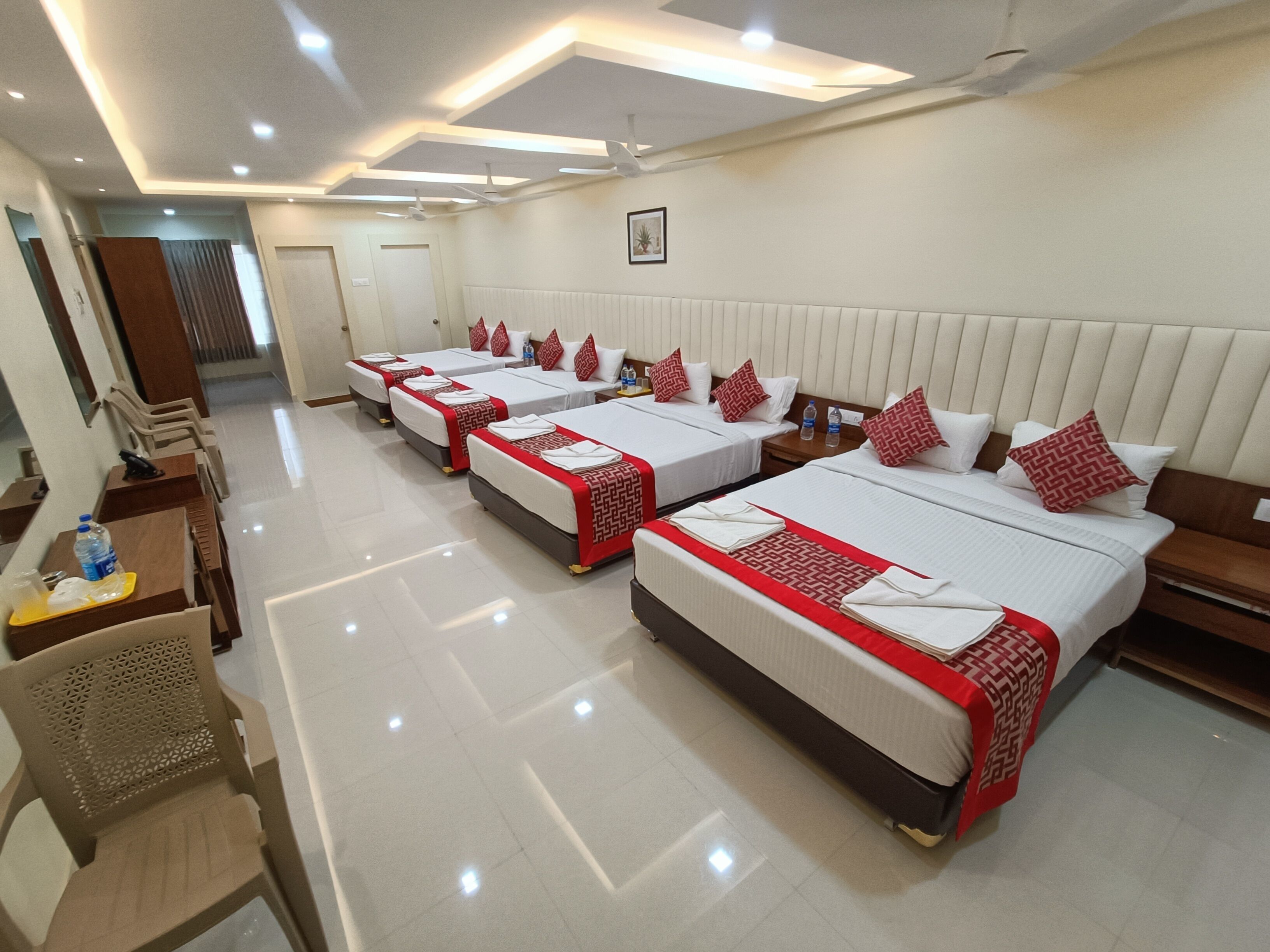 Une chambre d’hôtel avec quatre lits alignés, chacun recouvert d’une courtepointe à motifs rouges et blancs, et agrémenté d’oreillers assortis. Il y a des tables de chevet avec des bouteilles d’eau et un tableau encadré accroché au mur au-dessus des lits.