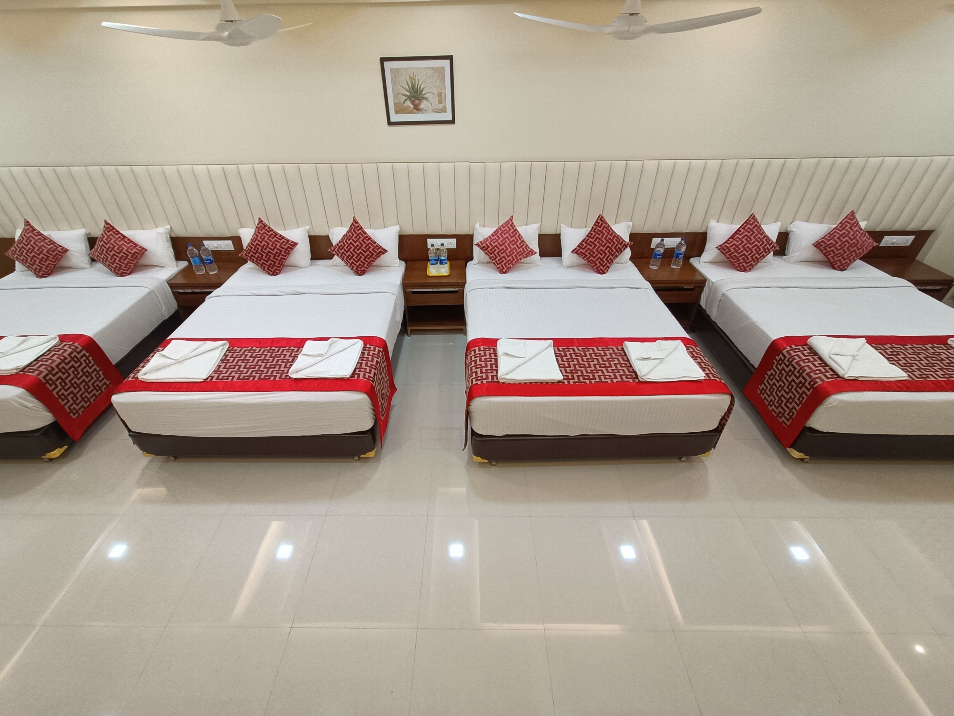 A hotel room with four beds arranged in a row, each with a red and white patterned bedspread, white pillows, and a small bedside table with bottled water.