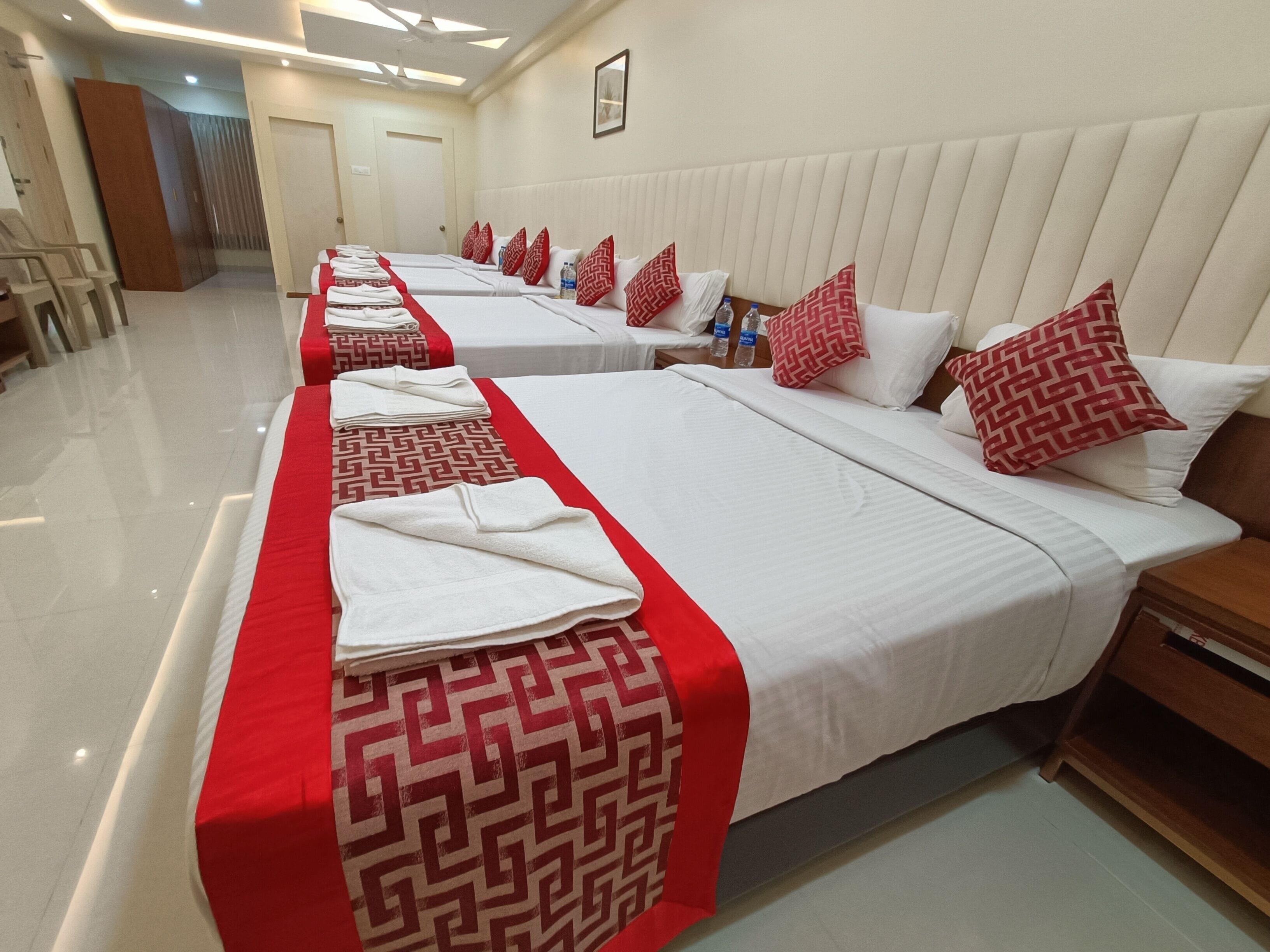 Quarto de hotel com duas camas, cada uma com colcha estampada em vermelho e branco, travesseiros brancos e almofada geométrica em vermelho e branco. Há duas mesas de cabeceira de madeira com garrafas de água sobre elas.