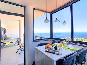 Un coin repas avec une table dressée pour deux personnes, une vue sur l’océan et un fauteuil suspendu en arrière-plan.