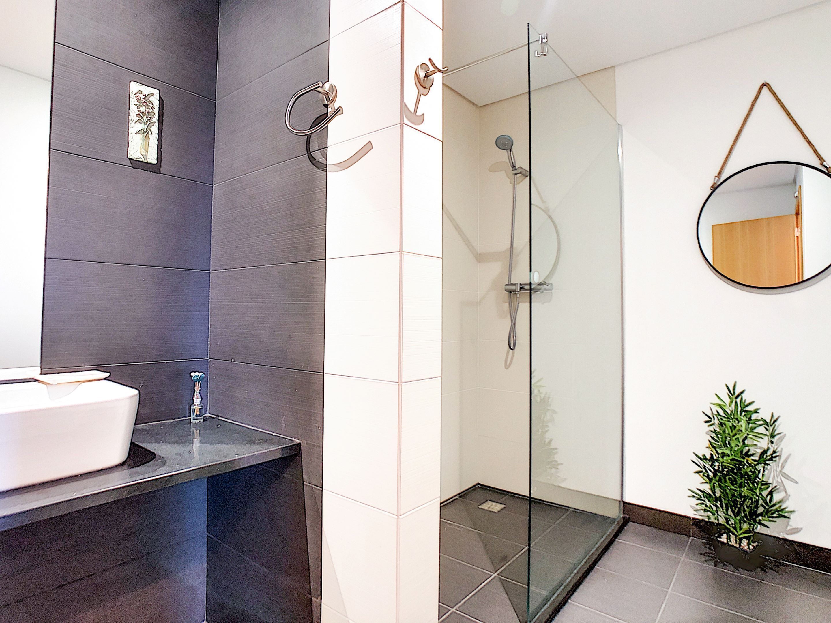A modern bathroom with a glass-enclosed shower, a round mirror, and a potted plant.