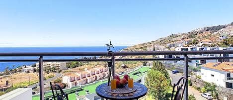 A balcony with a table and chairs, overlooking a coastal town with buildings and a clear blue sky.
