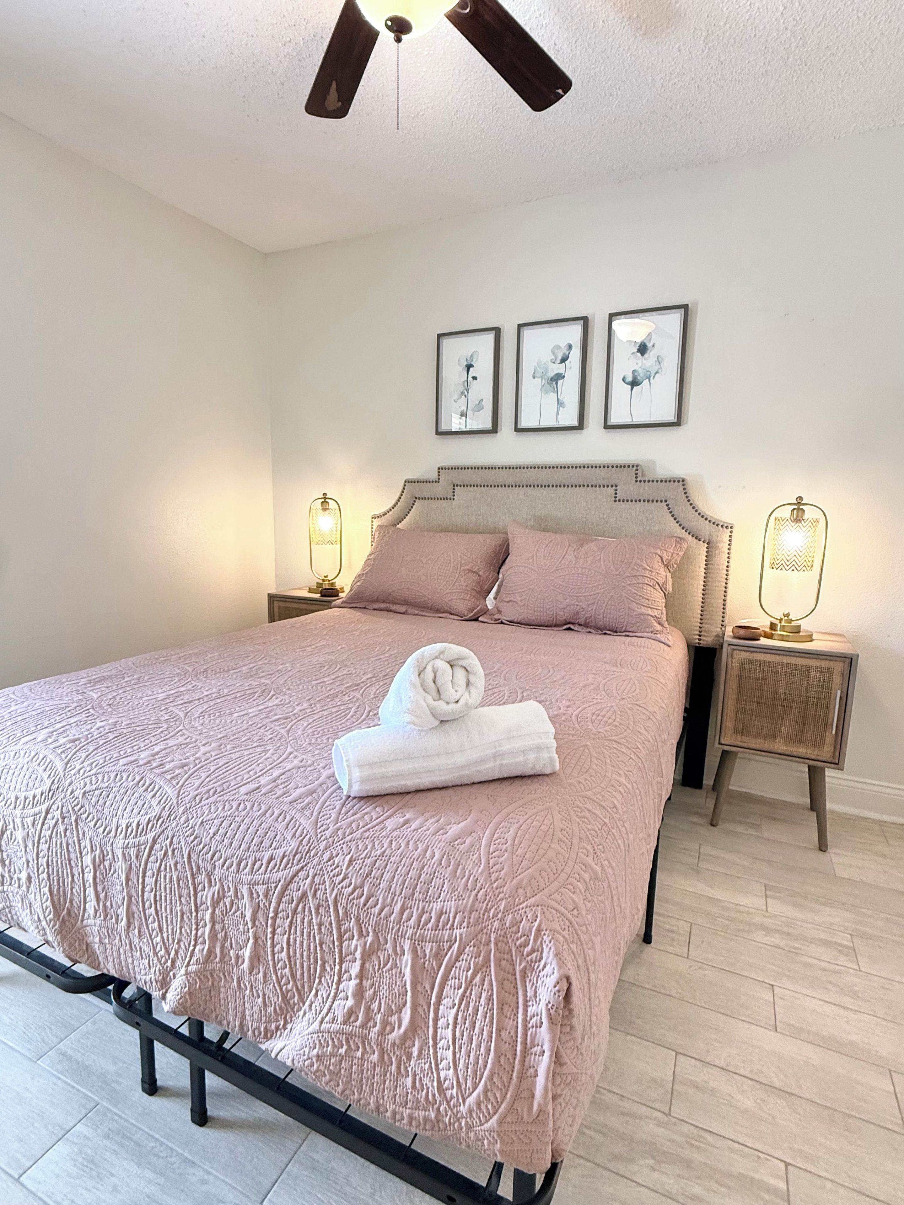 A neatly made bed with a patterned bedspread, two pillows, and a rolled towel on the bed. There are two bedside tables with lamps, and the room has a ceiling fan.