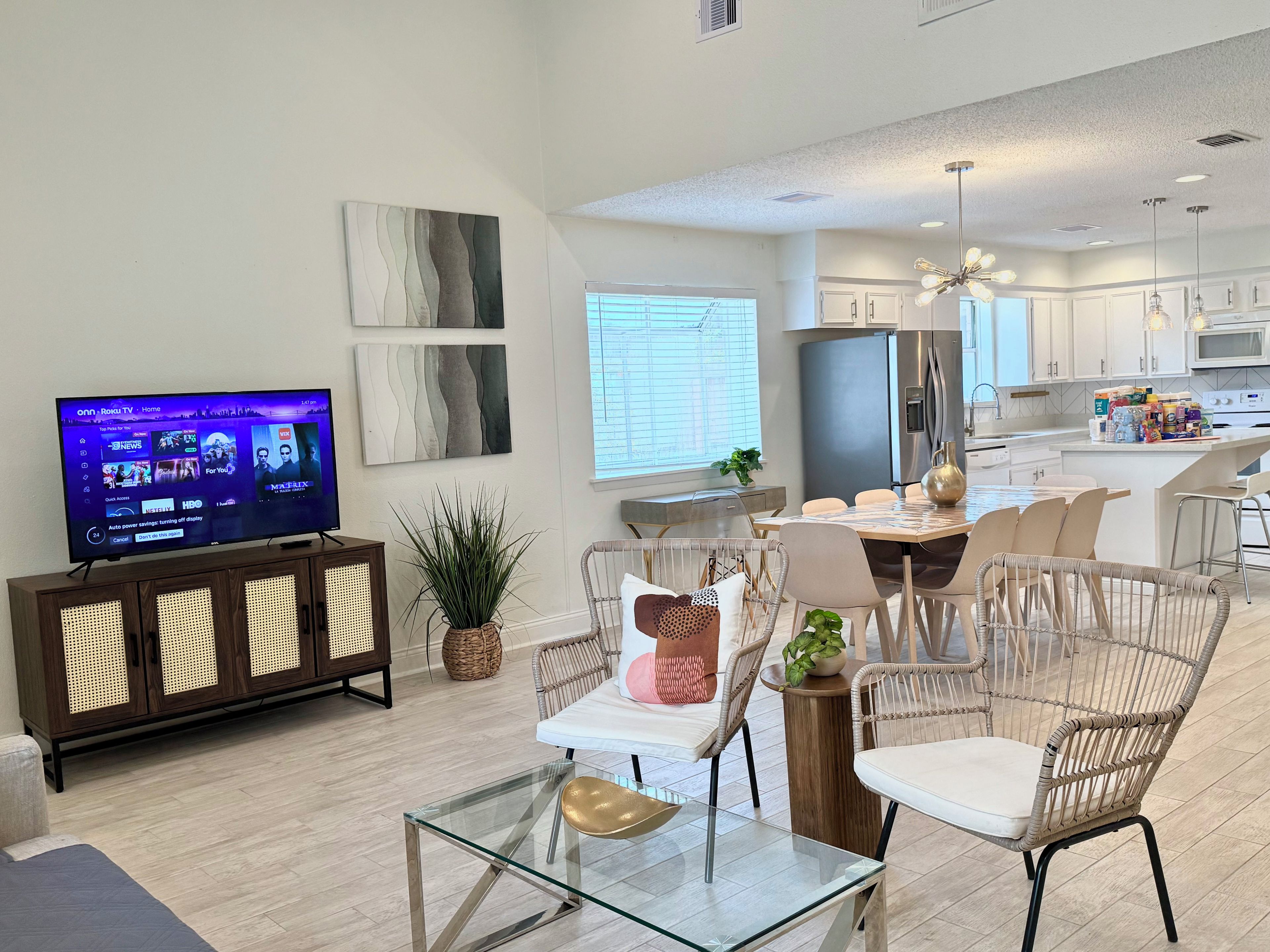 A modern living room with a flat-screen TV, wicker chairs, a glass coffee table, and a dining area with a white table and chairs.