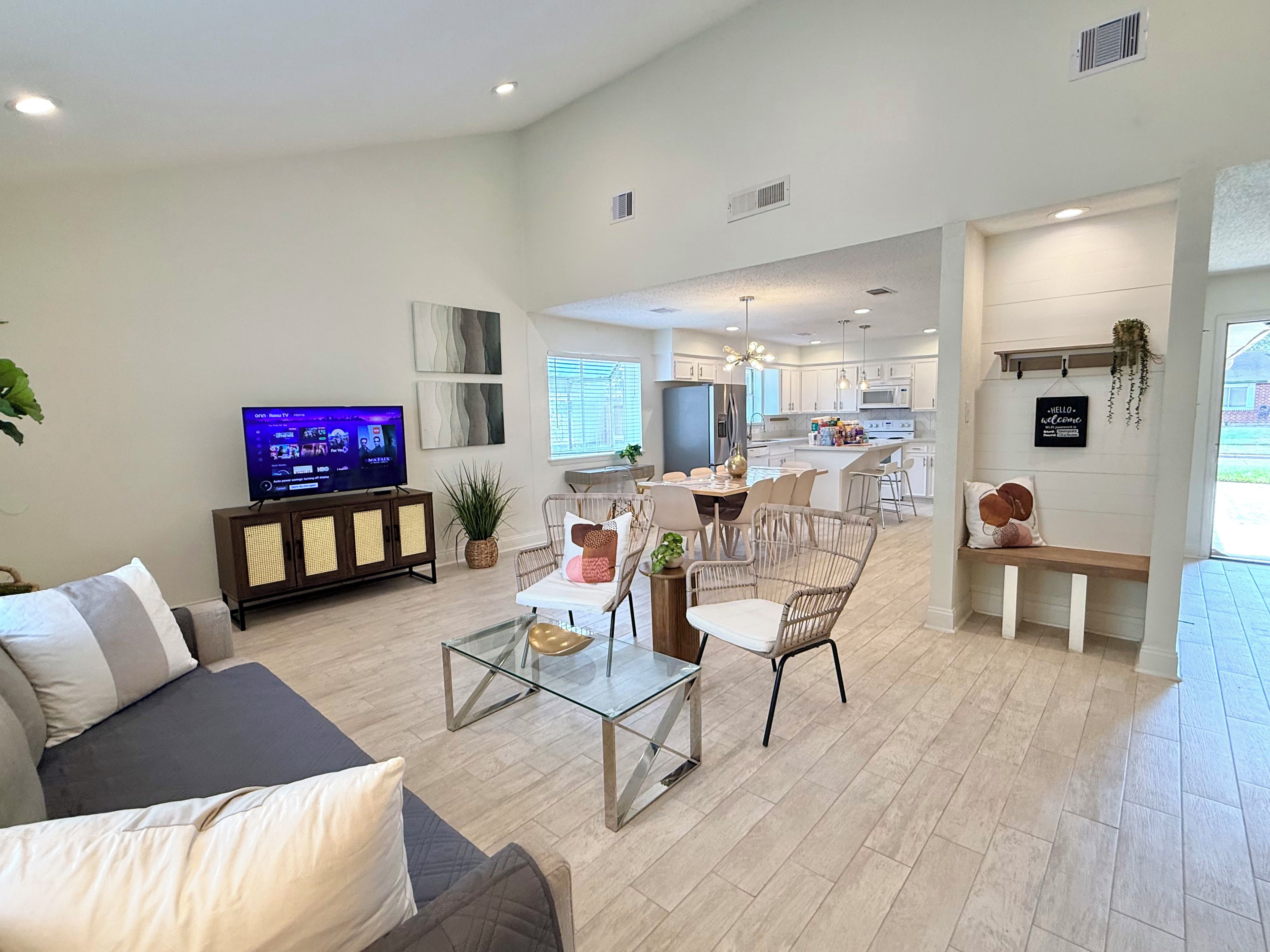 A modern living room with a flat-screen TV, a glass coffee table, and a dining area with wicker chairs.