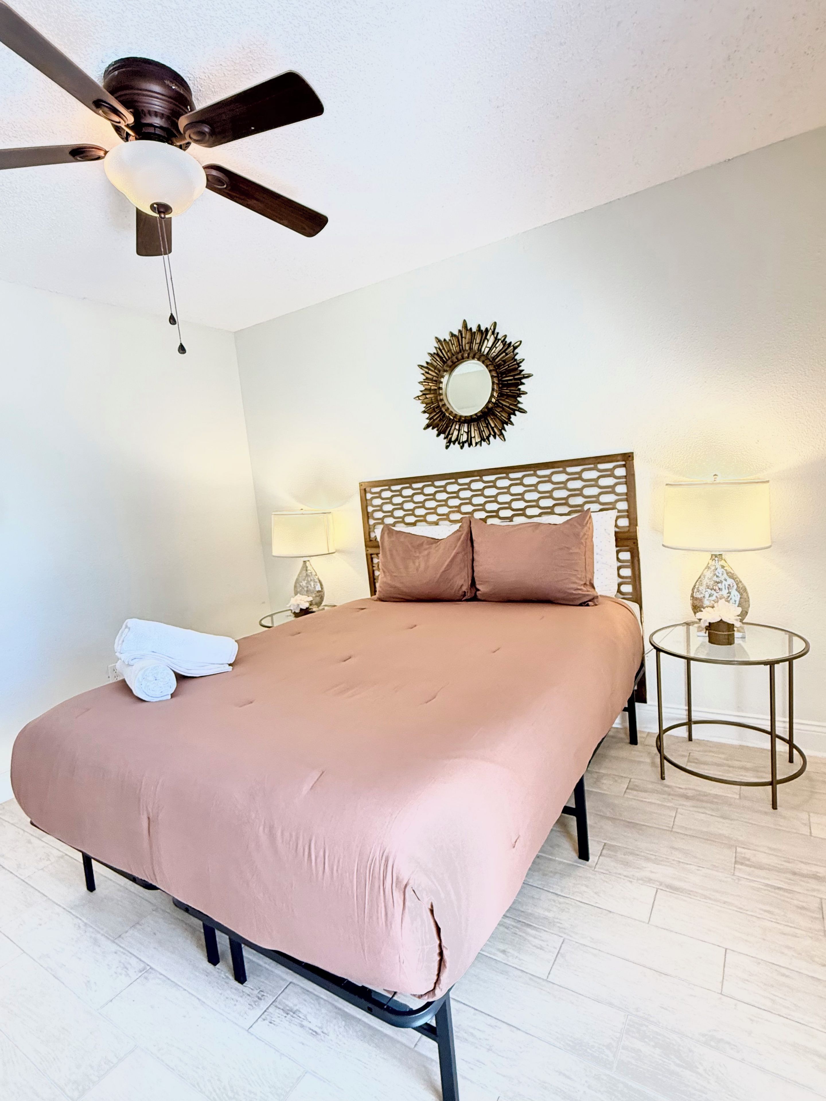 Une chambre à coucher comprenant un lit, un ventilateur de plafond, deux lampes de chevet, un miroir décoratif et une petite table ronde.